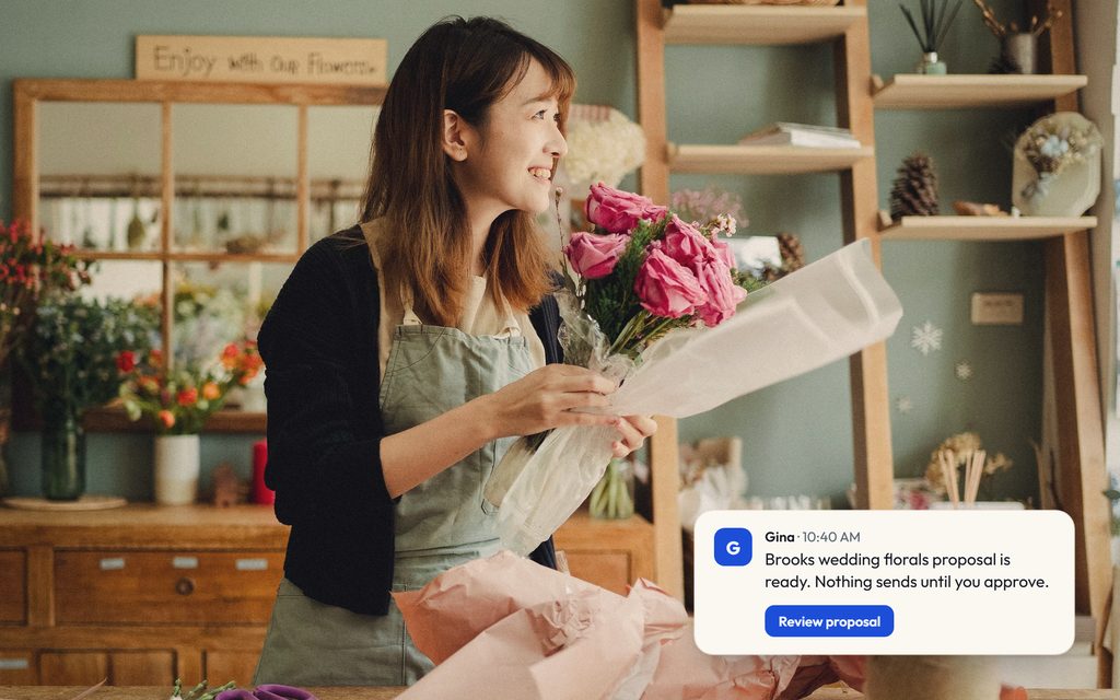 Florist wrapping a bouquet of pink roses at the shop bench, with a Gina message saying a florals proposal is ready for approval.
