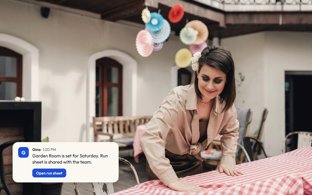Venue coordinator smoothing a tablecloth in a courtyard dressed for a party, with a Gina message confirming the room setup and run sheet.