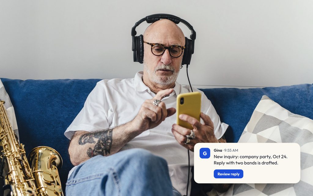 Booking agent in headphones reading a phone on the sofa beside a saxophone, with a Gina message about a new inquiry and a drafted reply.