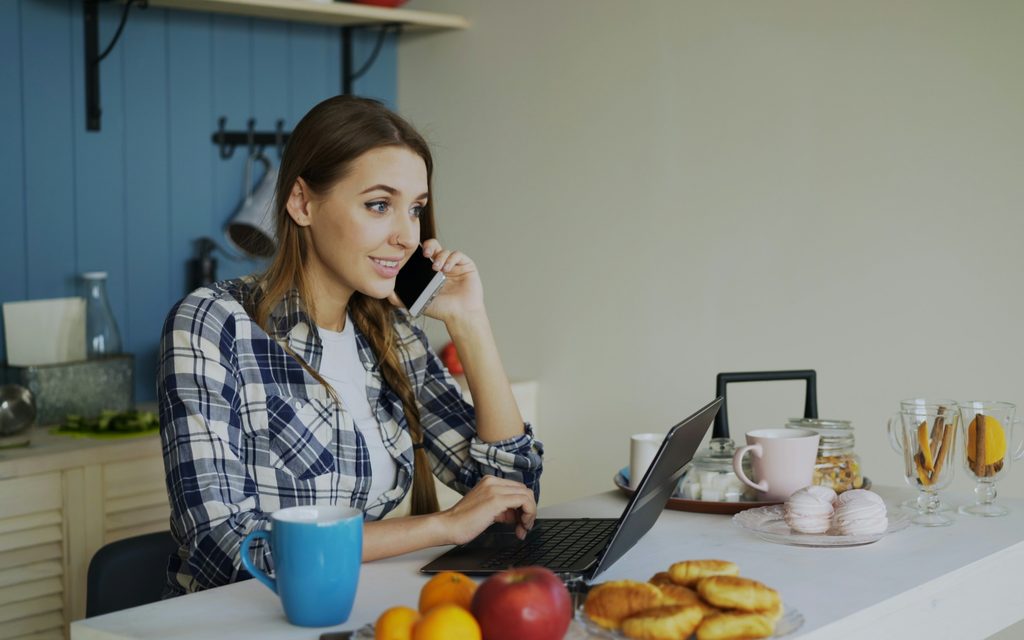 A woman at a sunlit kitchen table, on a call and smiling while she works on her laptop, with coffee and breakfast beside her.
