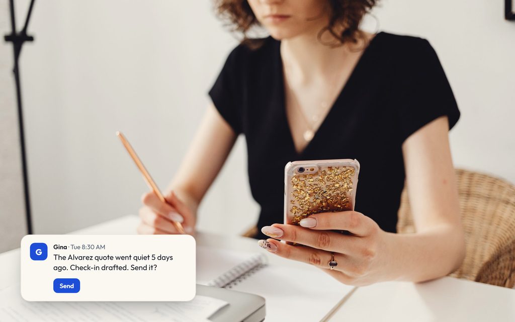 Planner checking a phone while jotting notes at a desk between setups, with a Gina message offering a drafted check-in on a quiet quote.